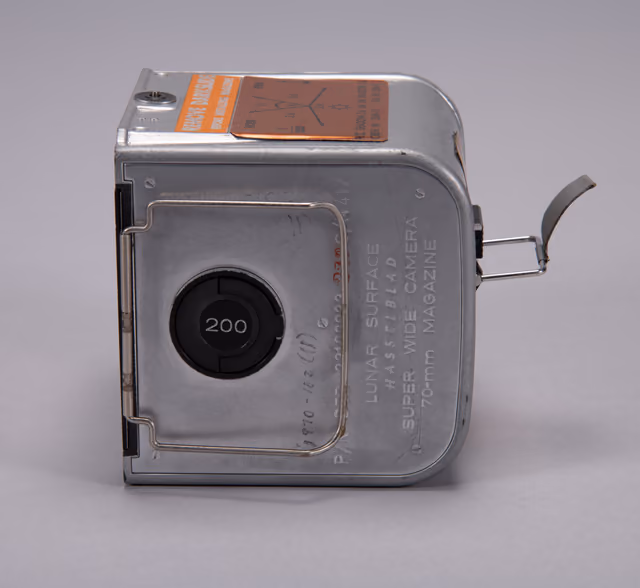 A 70mm Hasselblad film magazine used on the lunar surface during Apollo 11, a rectangular metal cassette photographed in museum lighting.